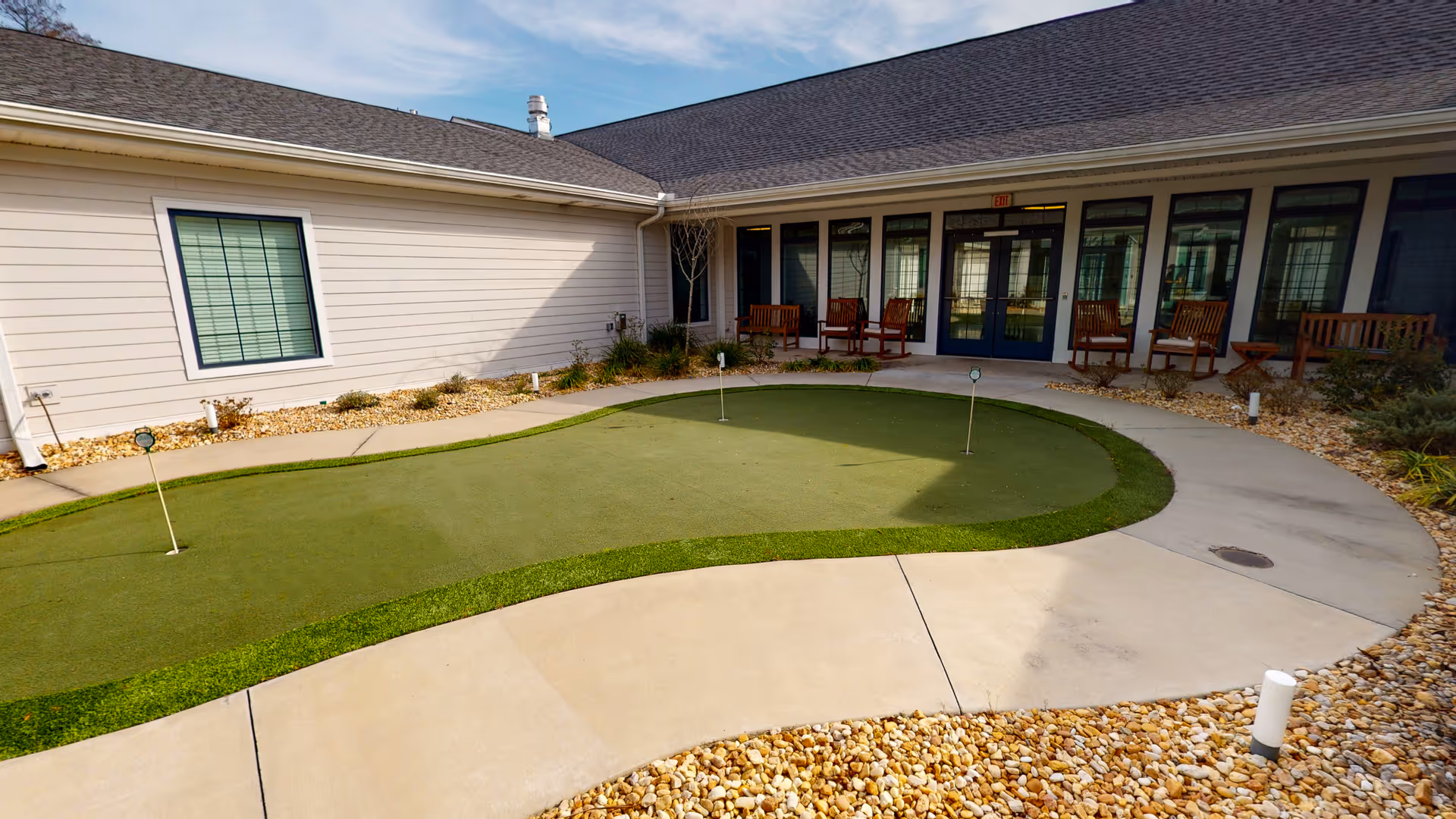 Courtyard with a small putting green and surrounding walkway in front of a single-story building with patio chairs.