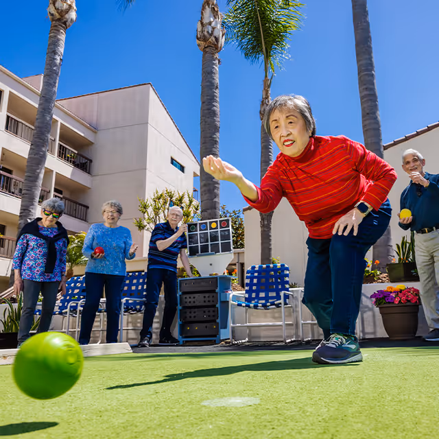 A group of elderly people playing lawn bowling outdoors on a sunny day at a senior living facility. One woman in a red striped sweater is in the foreground rolling a green ball, while others watch and hold balls in the background. Palm trees and a multi-story building are visible behind them.