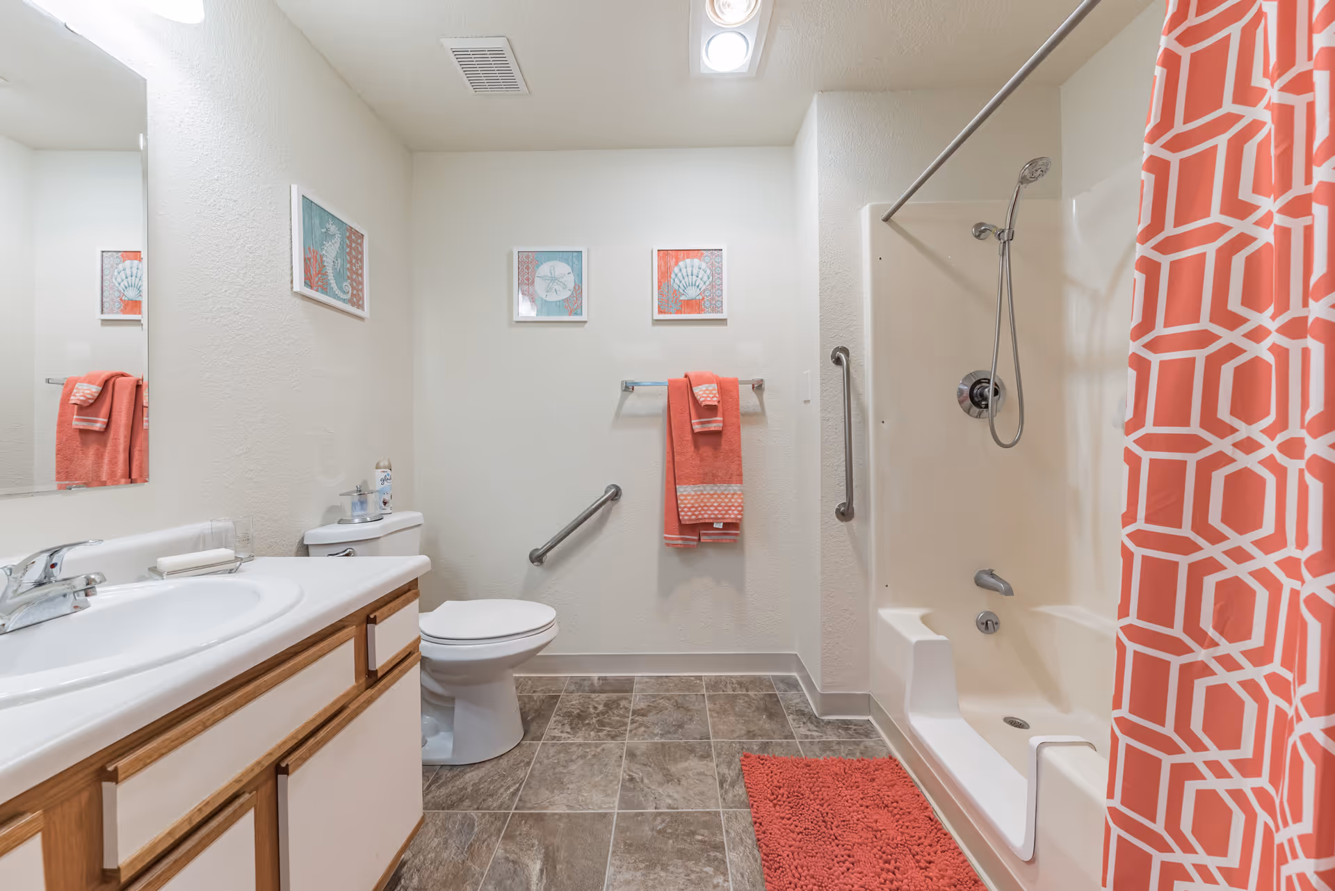 A clean bathroom with a white sink and wooden cabinet, a toilet with a can of air freshener on top, a bathtub with a handheld showerhead and a coral patterned shower curtain, coral towels hanging on a towel rack, coral bath mat on the floor, and three framed sea-themed pictures on the wall.