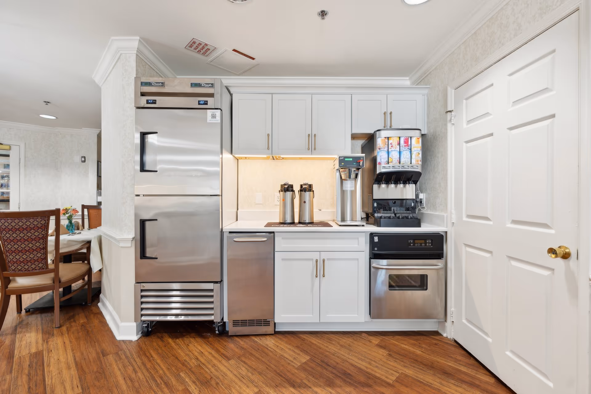 Small kitchenette with stainless steel refrigerator and appliances, white cabinets, and a beverage station beside a dining area.