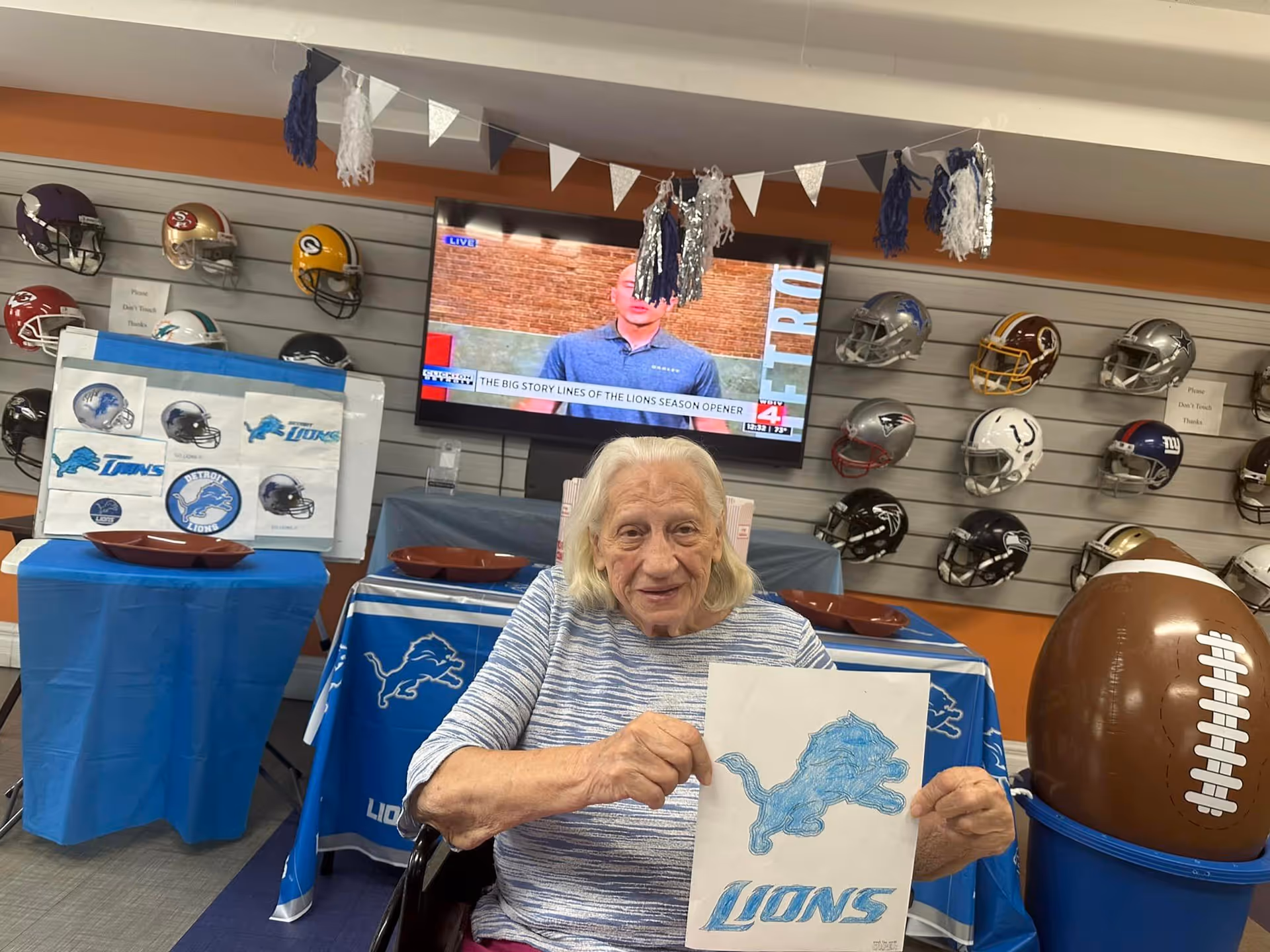 An elderly woman sits in a decorated indoor common room holding a colored Detroit Lions drawing, with football helmets on display and a TV mounted on the wall behind her.