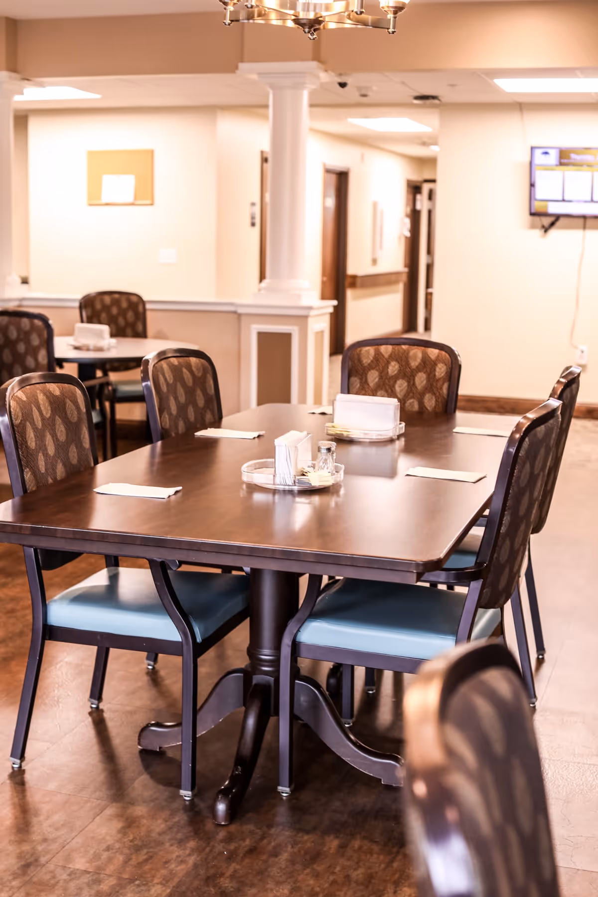 Interior view of a dining area with a dark wooden table and six chairs with patterned backs and blue seats. The table is set with napkins and condiments. The room has beige walls, a column, and a wall-mounted TV in the background.