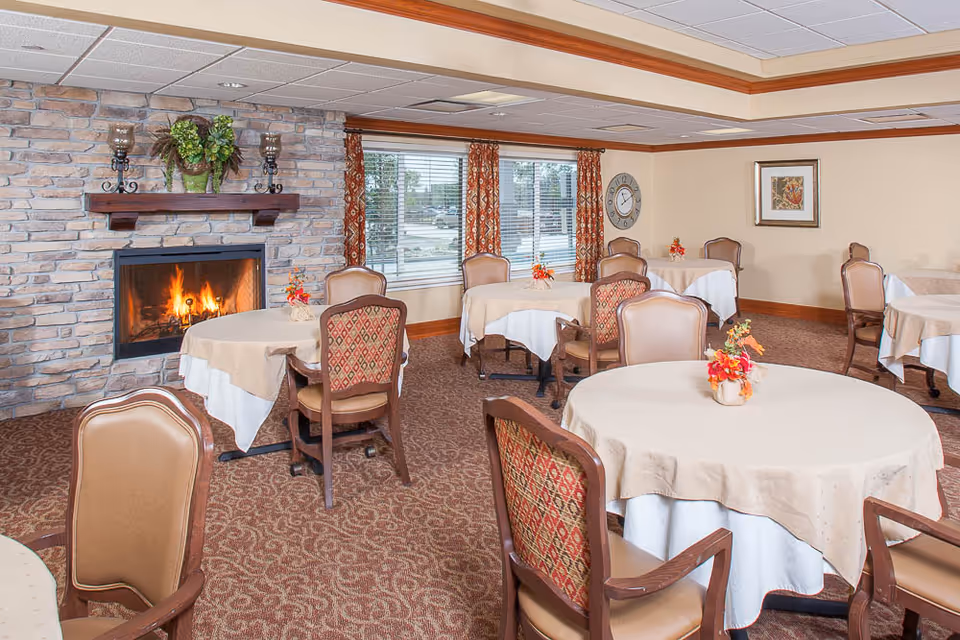 Dining room with round tables covered in tablecloths, upholstered chairs, floral centerpieces, and a lit fireplace set into a stone wall.