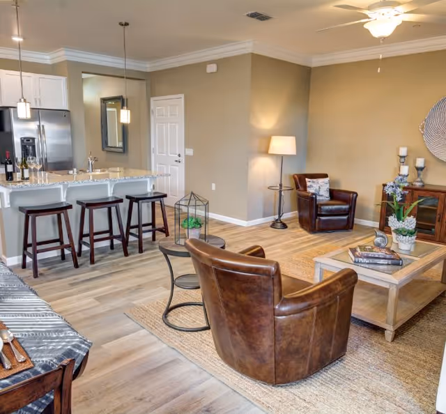 A cozy living room area with two brown leather armchairs, a wooden coffee table with books and a plant, a floor lamp, and a wooden cabinet with decorative items. The room has light wood flooring and beige walls. Adjacent to the living room is a kitchen area with a granite countertop island, three dark wooden bar stools, stainless steel refrigerator, and pendant lights hanging above the island.