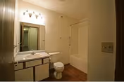 A small bathroom with a white sink and vanity, a toilet, and a bathtub with a shower curtain. The walls are light-colored, and there is a mirror above the sink with three light fixtures mounted above it. The floor appears to be wood or wood-like material.