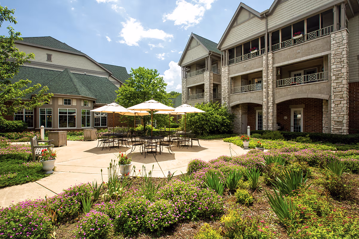 Outdoor patio area at Newcastle Place featuring several tables with umbrellas surrounded by landscaped flower beds and greenery, with a multi-story building in the background under a partly cloudy sky.