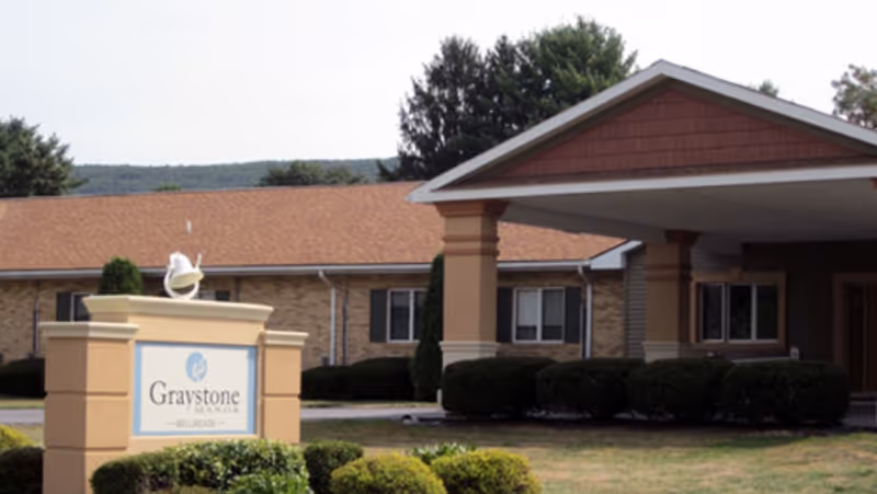 Exterior view of Graystone Manor Bellmeade senior living facility showing the building entrance with a covered driveway and a sign with the facility name in front surrounded by bushes and greenery.