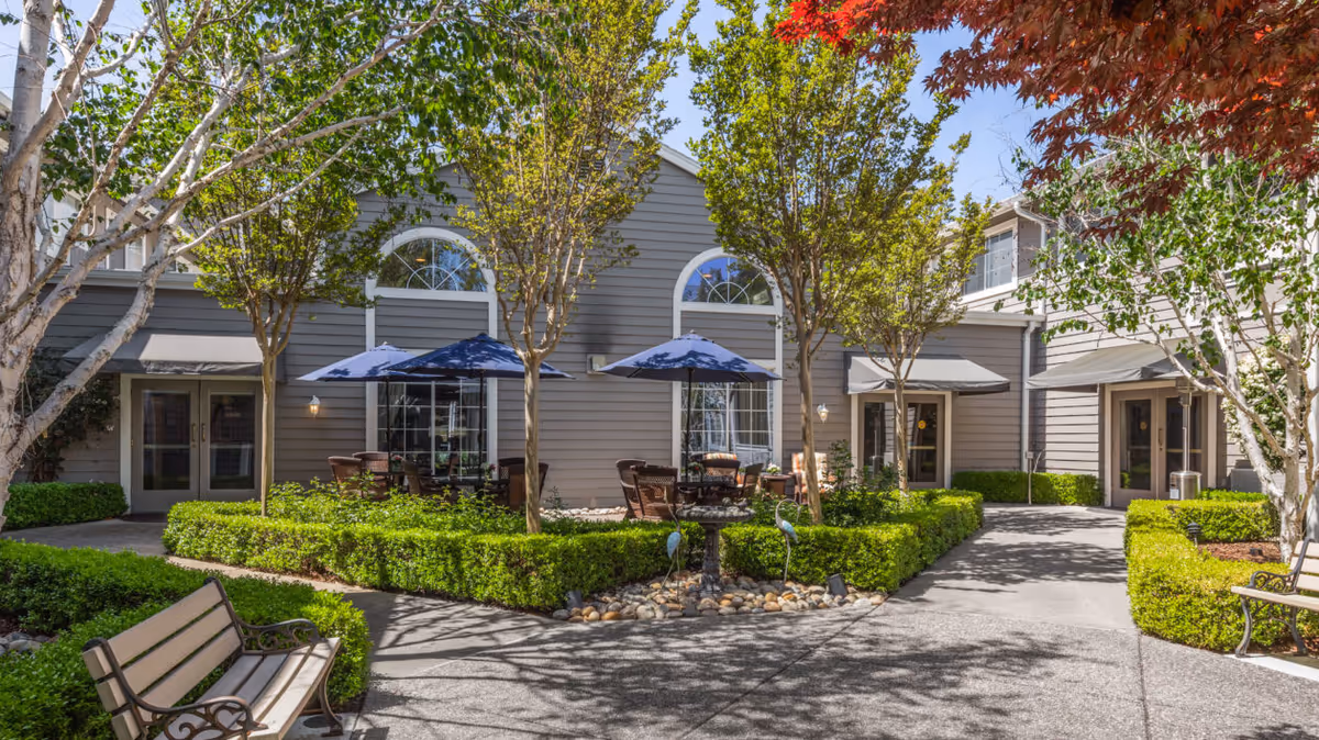 Outdoor courtyard area at North Shore Place featuring a paved walkway, green hedges, several trees, benches, and patio tables with blue umbrellas in front of a gray building with large arched windows.