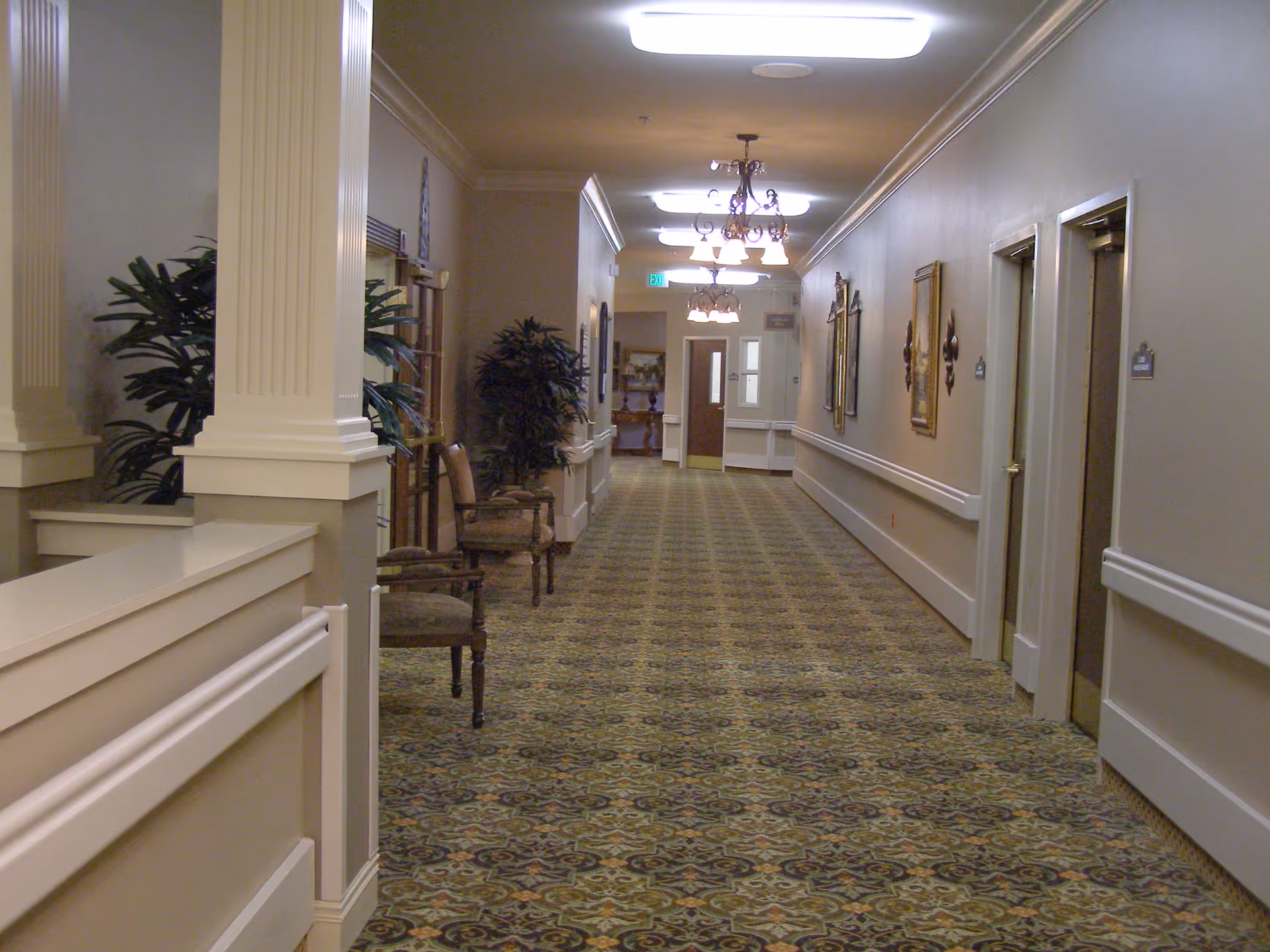 Long carpeted hallway in a senior care facility with chairs, potted plants, framed artwork, and overhead chandeliers.