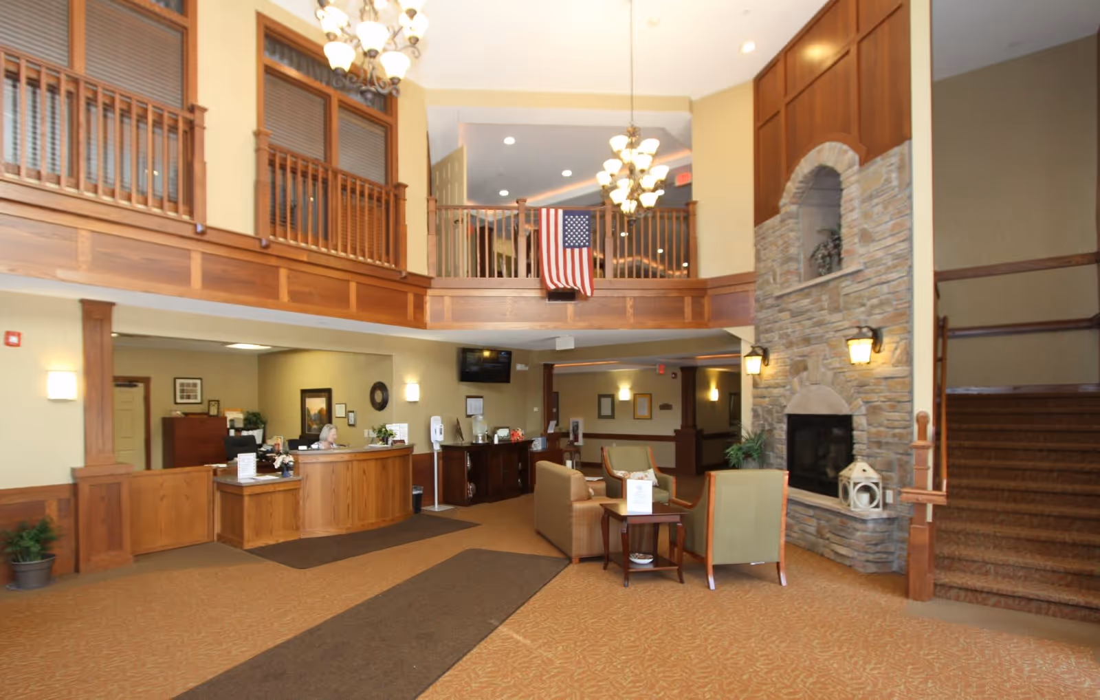 Interior view of a senior living facility lobby with a wooden reception desk on the left, a seating area with chairs and a small table in the center, a stone fireplace on the right, and a staircase leading upstairs. The upper level has wooden railings and an American flag hanging from it. The space is warmly lit with chandeliers and wall sconces.
