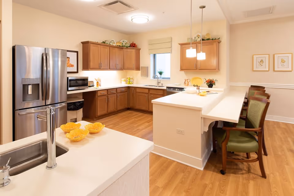 A bright and clean kitchen area with wooden cabinets, a stainless steel refrigerator, a microwave, and a sink with a modern faucet. There is a white countertop island with yellow decorative bowls and a breakfast bar with green cushioned chairs. The room has wooden flooring and soft lighting from ceiling fixtures and pendant lights.