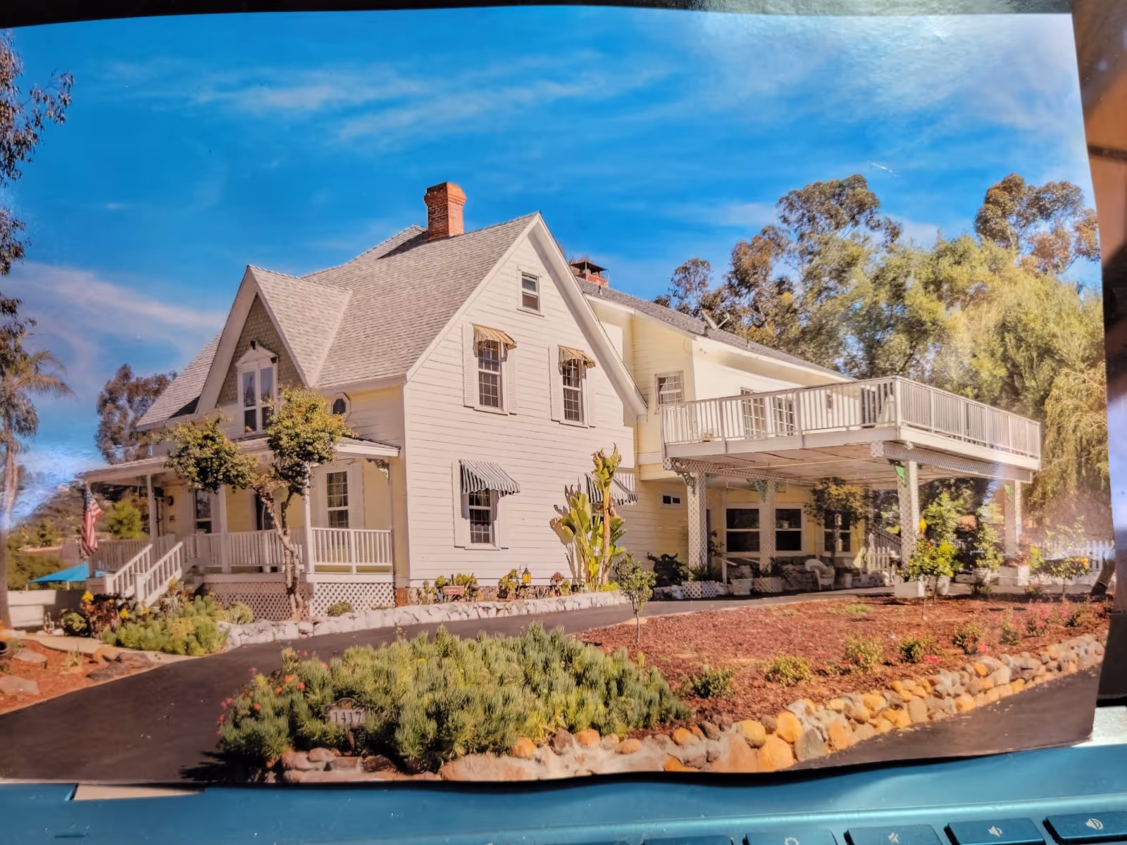 Large white two-story house with a wraparound porch and elevated deck surrounded by landscaping under a blue sky.