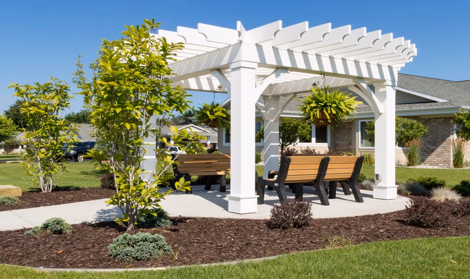 Outdoor seating area with wooden benches under a white pergola surrounded by landscaped garden beds with small trees and shrubs, adjacent to a brick building under a clear blue sky.