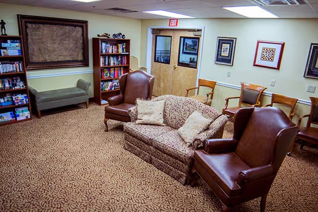 A cozy common area in an assisted living facility featuring a patterned carpet, a floral upholstered sofa with two cushions, two brown leather armchairs, wooden chairs along the wall, bookshelves filled with books and games, framed artwork on the walls, and double doors with glass panels.