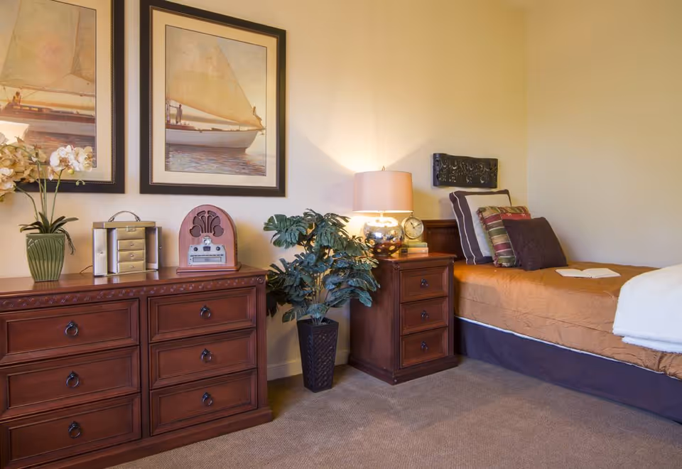 A cozy bedroom with a single bed covered in a brown quilt and multiple pillows. Next to the bed is a wooden nightstand with a lamp, clock, and books. A large wooden dresser with decorative items and two framed pictures of sailboats hang on the wall above it. A potted plant is placed between the dresser and the nightstand.