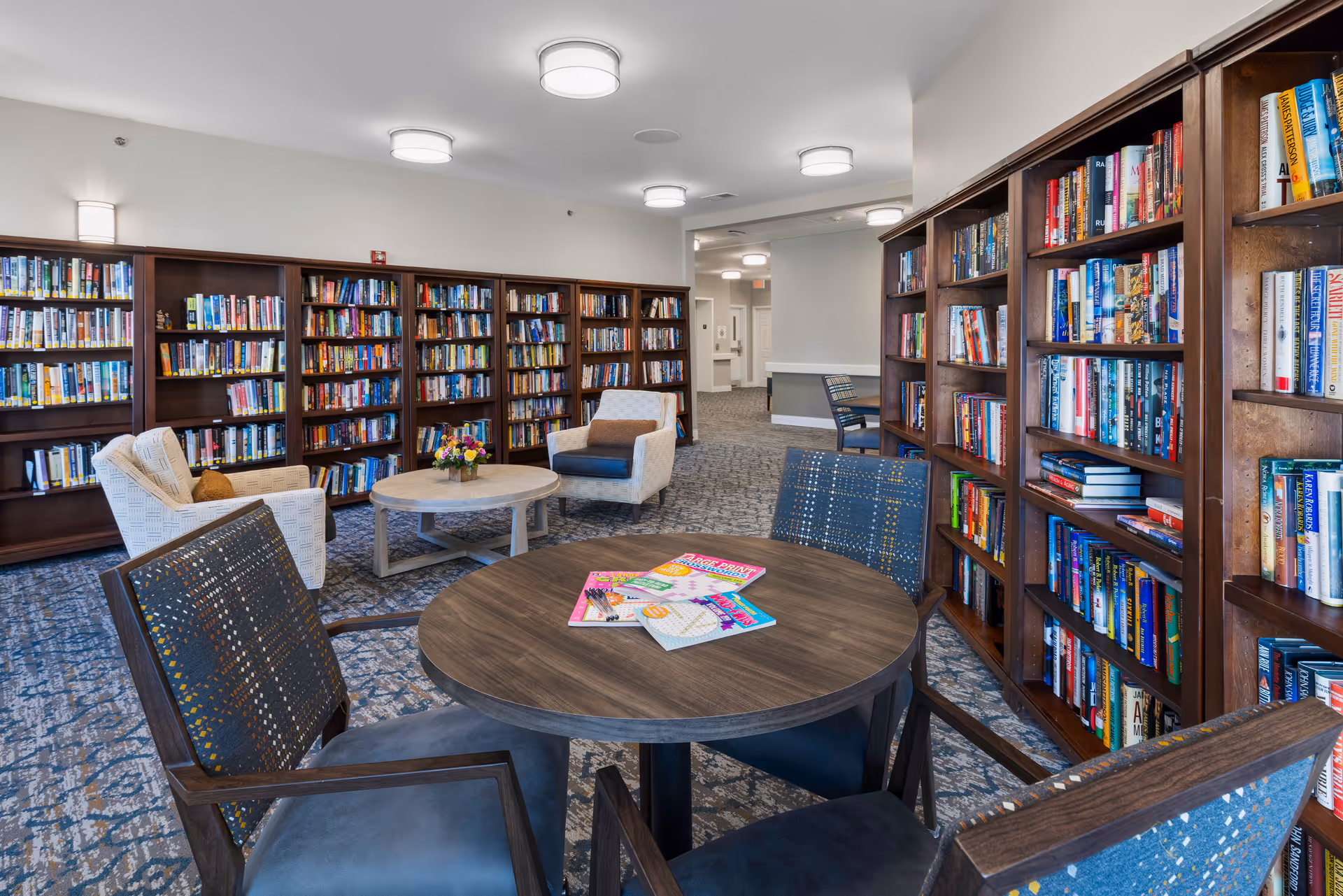 A bright communal library with bookshelves lining the walls and round tables with upholstered chairs arranged for reading.
