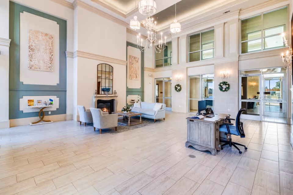 Spacious and elegant lobby area with high ceilings, chandeliers, a fireplace, and comfortable seating including a sofa and armchairs. There is a wooden desk with a computer and office chair near the entrance doors. The walls are decorated with artwork and wreaths, and large windows allow natural light to fill the space.