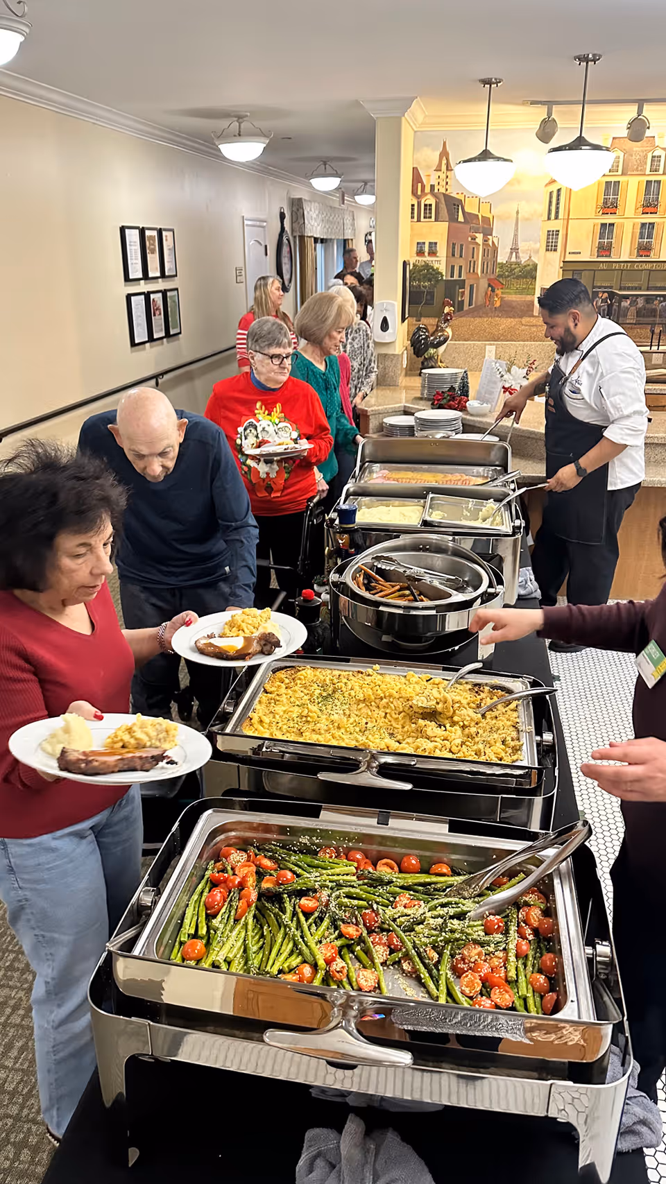 A group of elderly people serving themselves food from a buffet line in a dining area. The buffet includes dishes like roasted asparagus with cherry tomatoes, scrambled eggs, sausages, and other hot foods. A staff member in a white shirt and black apron is assisting at the end of the buffet. The background features a mural of a European street scene.