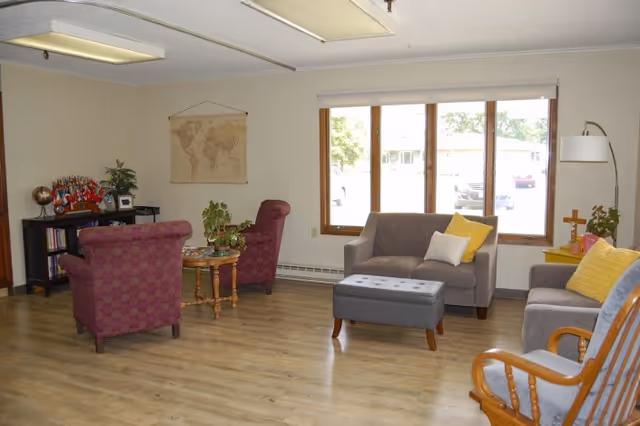 A cozy living room with wooden flooring, two purple armchairs, a gray loveseat with yellow and white pillows, a gray ottoman, a wooden rocking chair, and a small wooden table with plants. There is a large window letting in natural light and a world map hanging on the wall.