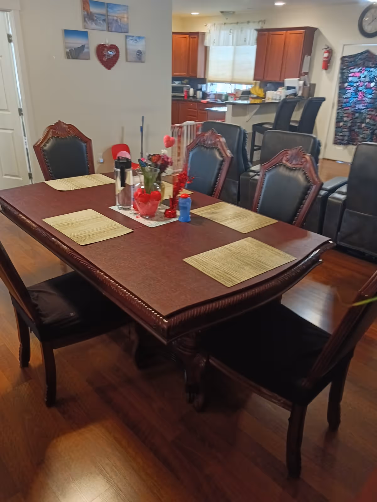 A dining area with a rectangular wooden table set with six chairs. The table has placemats, a water bottle, a vase with flowers, and small decorative items. In the background, there is a kitchen with wooden cabinets and a counter with bar stools. The walls have framed pictures and a heart-shaped decoration.
