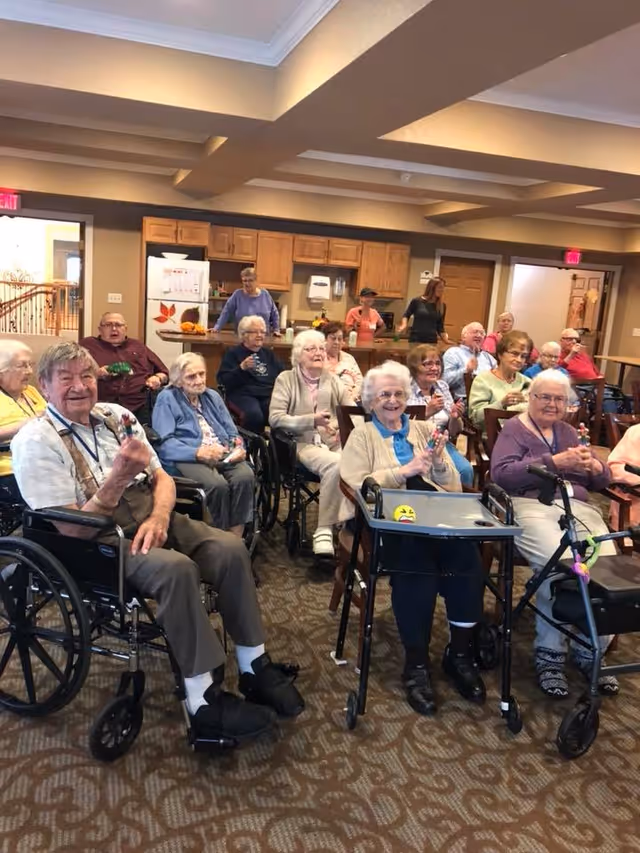 A group of elderly residents sitting in wheelchairs and chairs in a common room, smiling and holding small objects, with a kitchen area and staff members in the background.