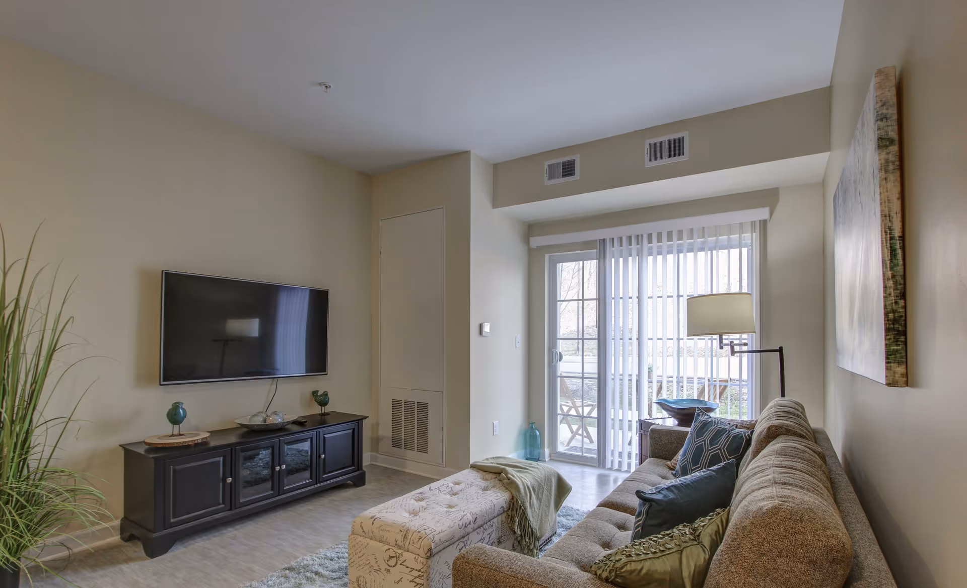 A cozy living room with a beige couch adorned with multiple pillows, a patterned ottoman with a green throw blanket, a black TV stand with decorative items, a wall-mounted flat-screen TV, a floor lamp, a large window with vertical blinds, and a sliding glass door leading to an outdoor patio with chairs.