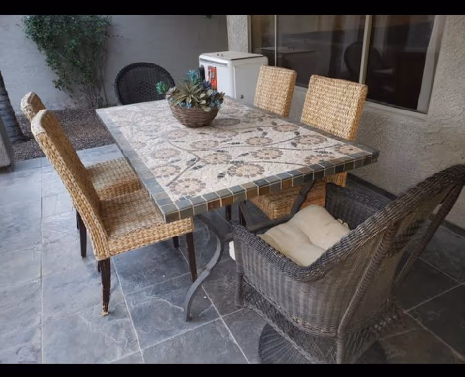 Outdoor patio area with a rectangular mosaic tile table surrounded by six wicker chairs, some with cushions. There is a potted succulent centerpiece on the table and a small white appliance against the wall in the background.