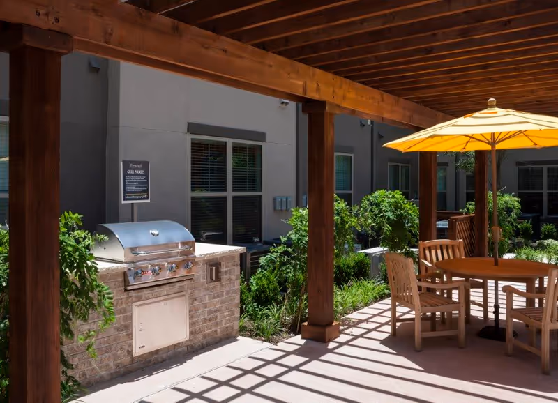 Outdoor patio area with a built-in stainless steel grill set in a brick counter, surrounded by greenery and plants. There is a wooden pergola overhead casting shadows on the ground. A round wooden table with four wooden chairs and a yellow umbrella is also visible.
