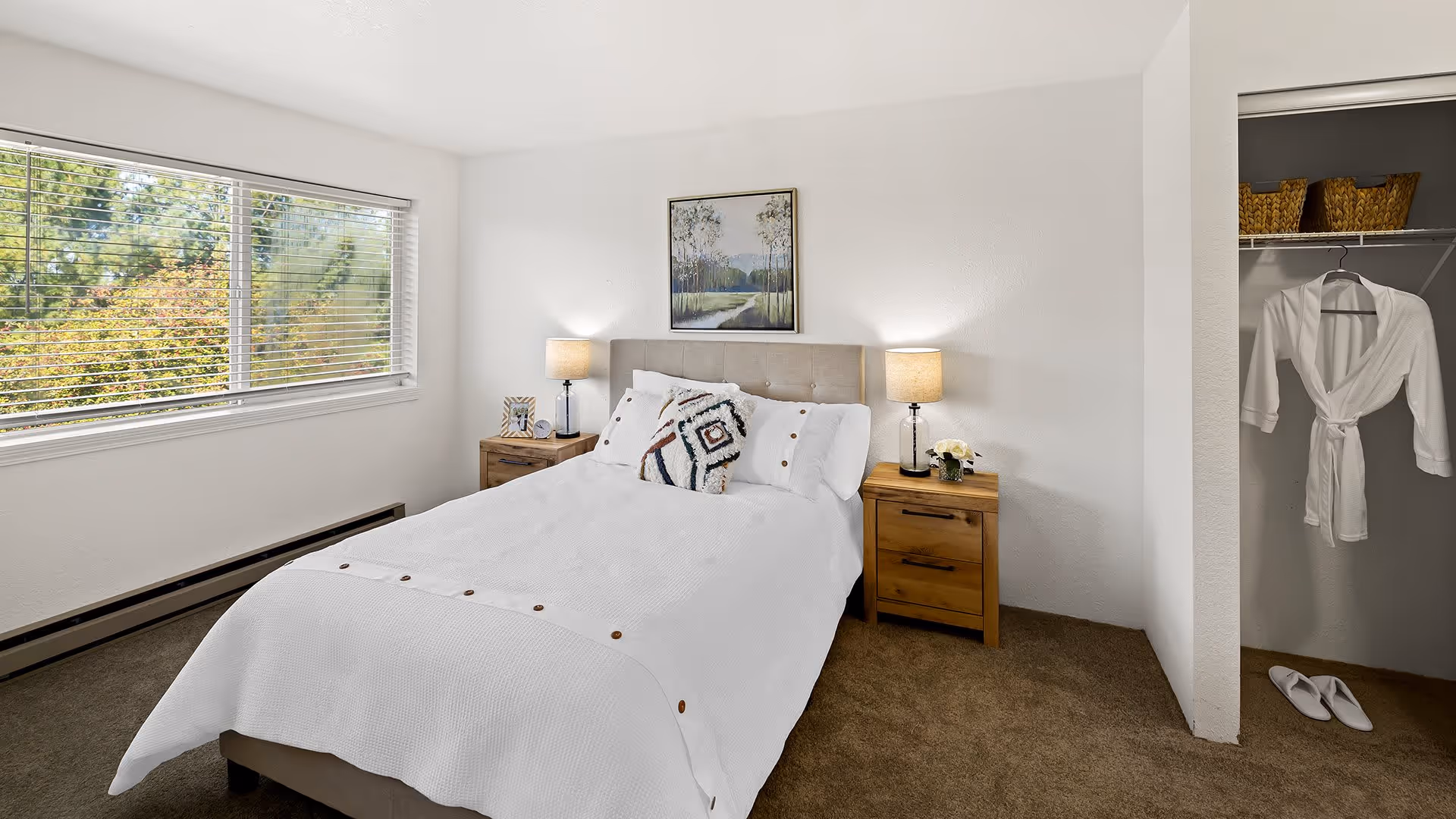A bright bedroom with a large window showing greenery outside. The room features a neatly made bed with white bedding and decorative pillows, two wooden nightstands with lamps, a framed landscape painting above the bed, and an open closet with a white robe and slippers.