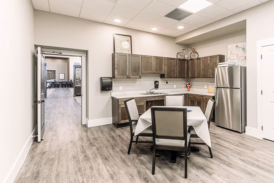 A clean and modern kitchen area in a senior living facility with wooden cabinets, a stainless steel refrigerator, a sink, and a small dining table covered with a white tablecloth surrounded by four chairs. The room has light-colored walls and wood-patterned flooring. An open door leads to a hallway with additional seating visible in the distance.