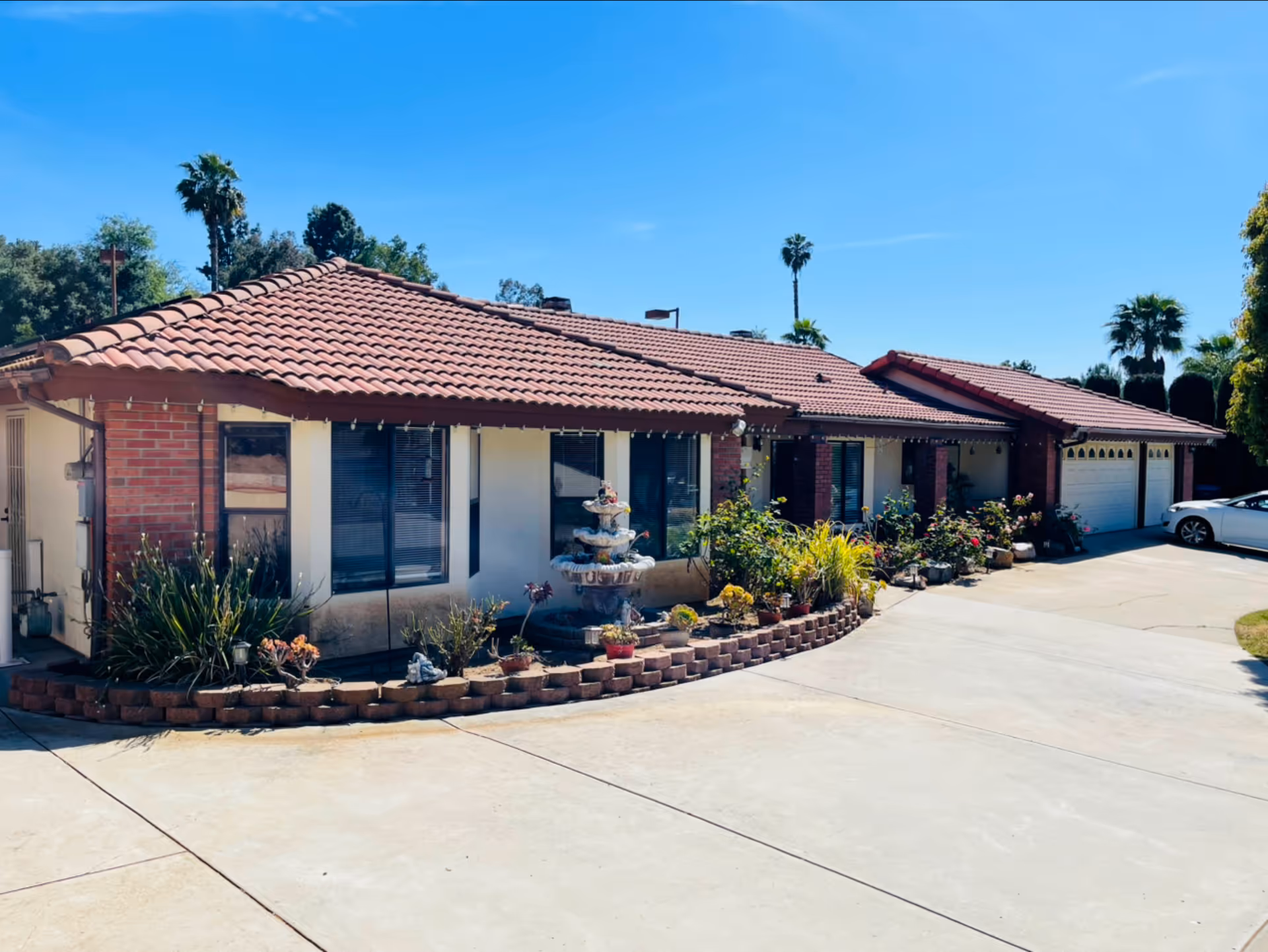 Exterior view of a single-story building with a red tile roof and brick accents, surrounded by plants and a small garden with a tiered fountain in front. A driveway leads to a three-car garage, and a white car is parked nearby. Tall palm trees and a clear blue sky are visible in the background.