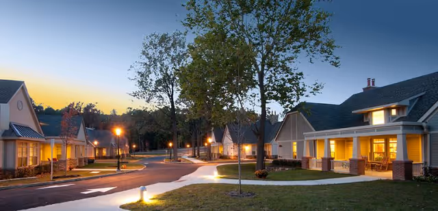 Evening view of a senior living community with single-story houses along a curved road, illuminated street lamps, and trees with some autumn foliage.