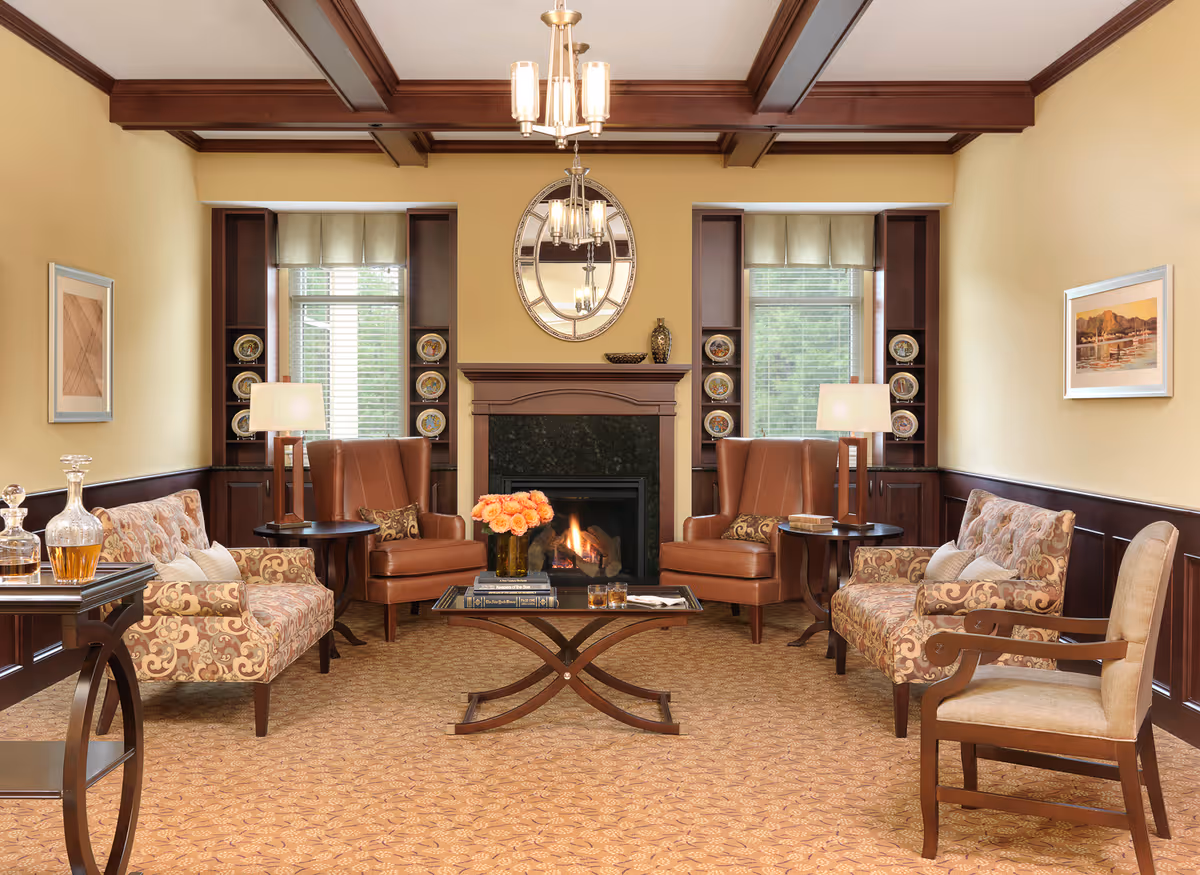 A cozy living room with a fireplace in the center, flanked by two windows with built-in shelves displaying decorative plates. The room features two brown leather armchairs, two patterned upholstered sofas, a beige armchair, and a glass coffee table with a vase of orange flowers and books. There are two table lamps on side tables next to the armchairs, a chandelier hanging from the ceiling, and framed artwork on the walls.