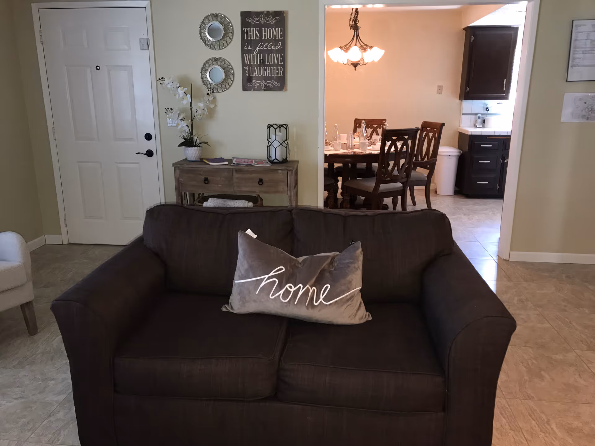 Interior view of a living room area with a dark brown couch featuring a decorative pillow with the word 'home'. Behind the couch is a small wooden table with a potted plant, a candle holder, and some magazines. On the wall above the table are two round mirrors and a sign that reads 'This home is filled with love & laughter'. To the right, a dining area with a wooden dining table and chairs is visible, set with glassware and plates. The kitchen area with dark cabinets and a white trash bin is seen further in the background.