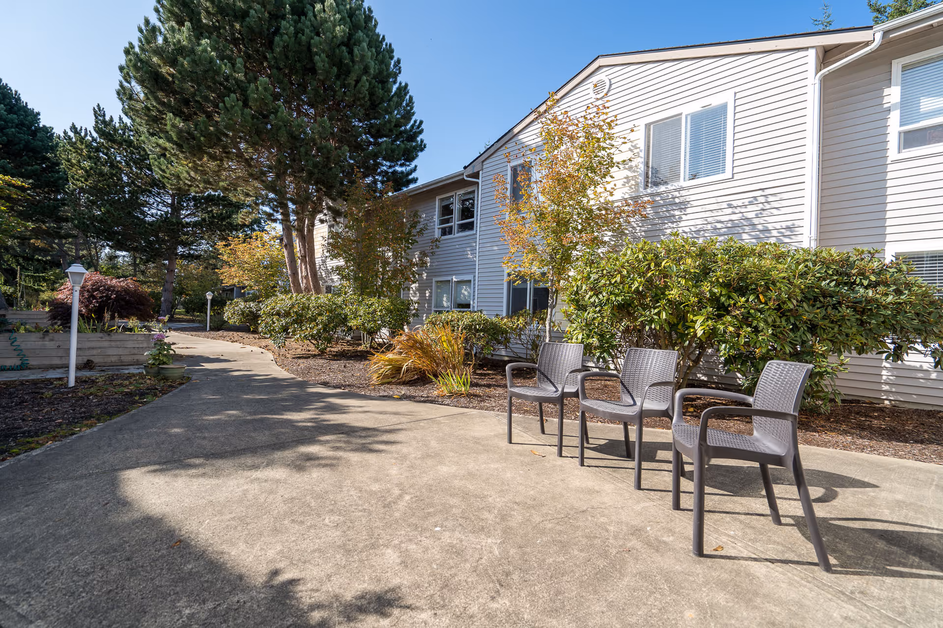 Outdoor paved walkway with three empty chairs along the side, surrounded by bushes and trees, next to a light-colored two-story building under a clear blue sky.