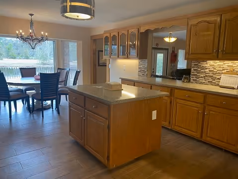 Open kitchen with a central island, wooden cabinets, granite countertops, and a dining area with a table and chandelier by large windows.