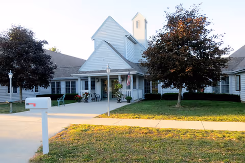 Exterior view of a senior living facility building with white siding, a peaked roof, and a small tower-like structure. There are two trees with dark foliage on either side of the entrance, a white mailbox in the foreground, and a concrete walkway leading to the entrance. An American flag is displayed near the door.