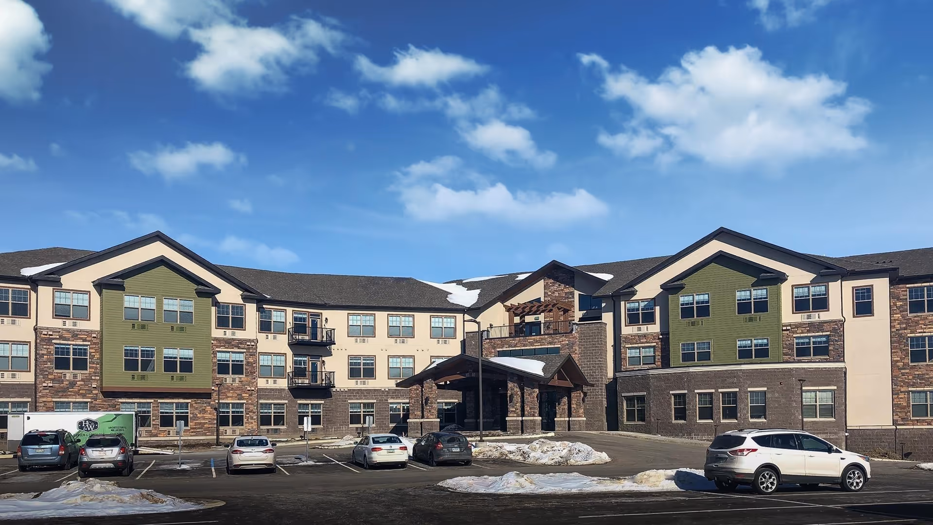 Exterior view of a three-story senior living facility building with a mix of beige, green, and stone facade under a blue sky with scattered clouds. Several cars are parked in the parking lot in front of the building, with patches of snow on the ground.