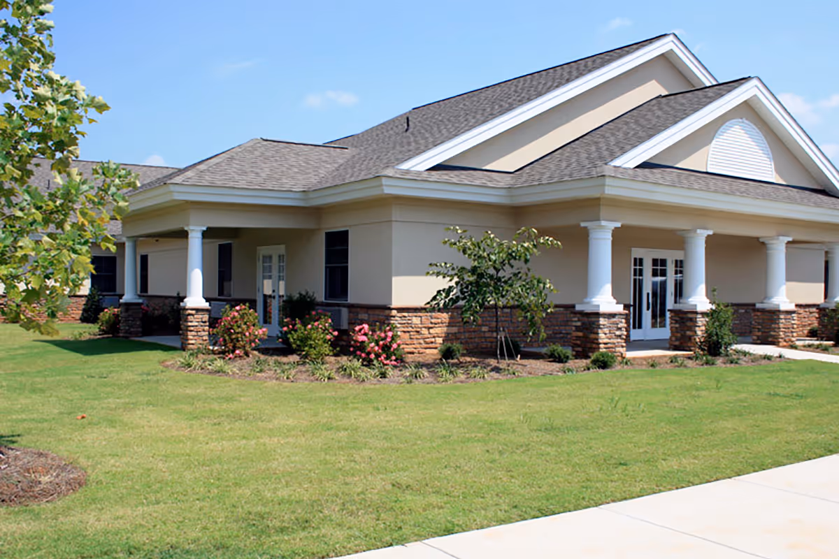 Exterior view of a single-story building with beige walls, white columns, and a gray shingled roof. The building is surrounded by a well-maintained lawn with small trees and flowering bushes under a clear blue sky.