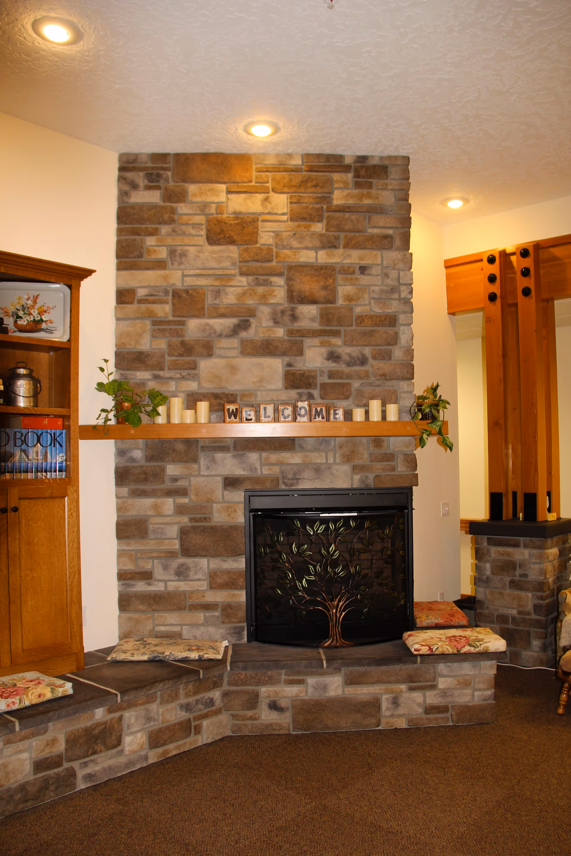 Cozy living room area featuring a stone fireplace with a decorative metal screen depicting a tree. Above the fireplace is a wooden mantel with candles, plants, and blocks spelling out 'WELCOME'. Cushioned stone seating surrounds the fireplace. To the left, there is a wooden cabinet with books and decorative items. The room has warm lighting from recessed ceiling lights.
