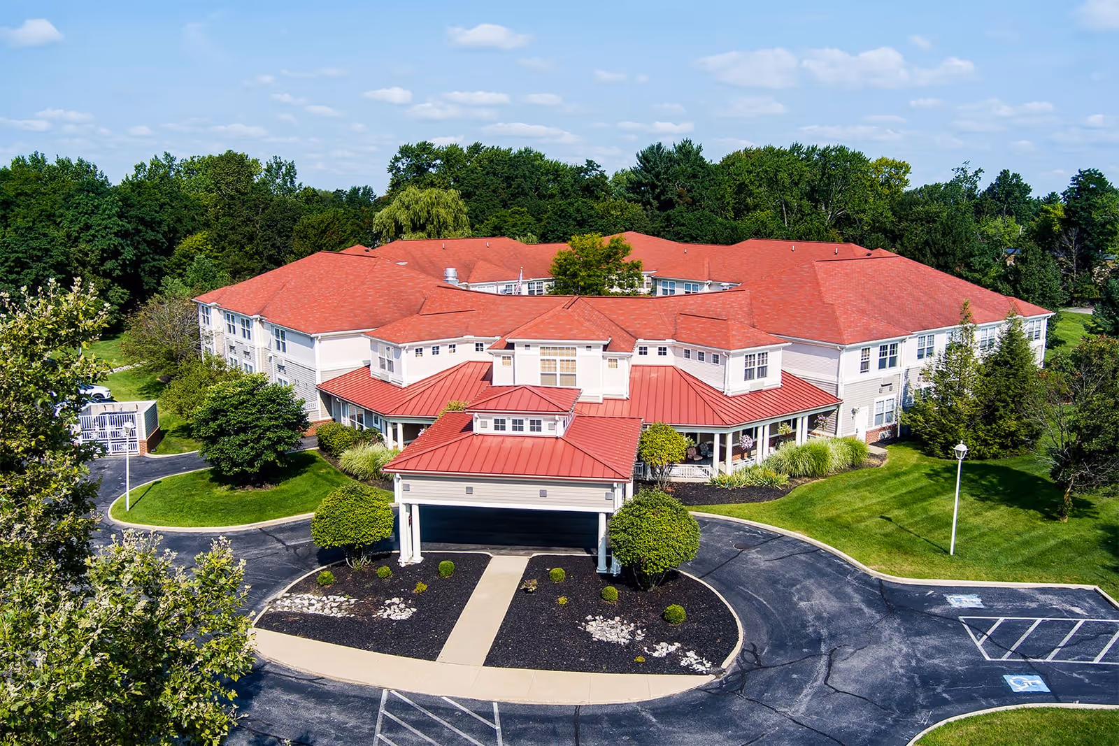 Aerial view of a large senior living facility with red roofs surrounded by green trees and landscaped lawns under a partly cloudy sky.