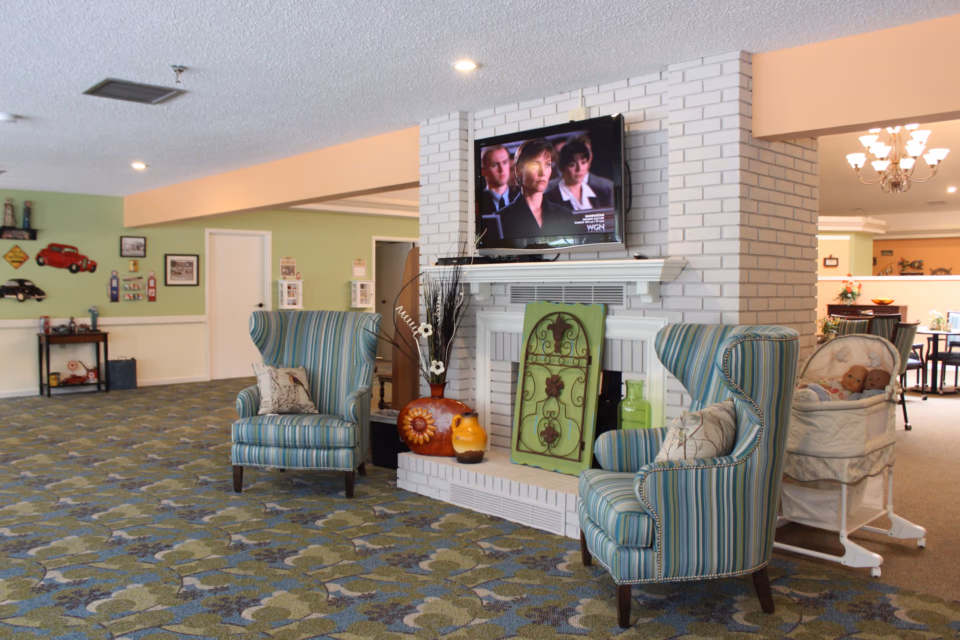 A cozy senior living common area with two striped armchairs facing a white brick fireplace. Above the fireplace is a mounted flat-screen TV showing a scene from a TV program. Decorative items including vases and a green ornamental panel are placed on the fireplace hearth. The room has patterned carpet flooring and light green walls with various wall decorations. In the background, there is a bassinet with two dolls and a dining area with tables and chairs.