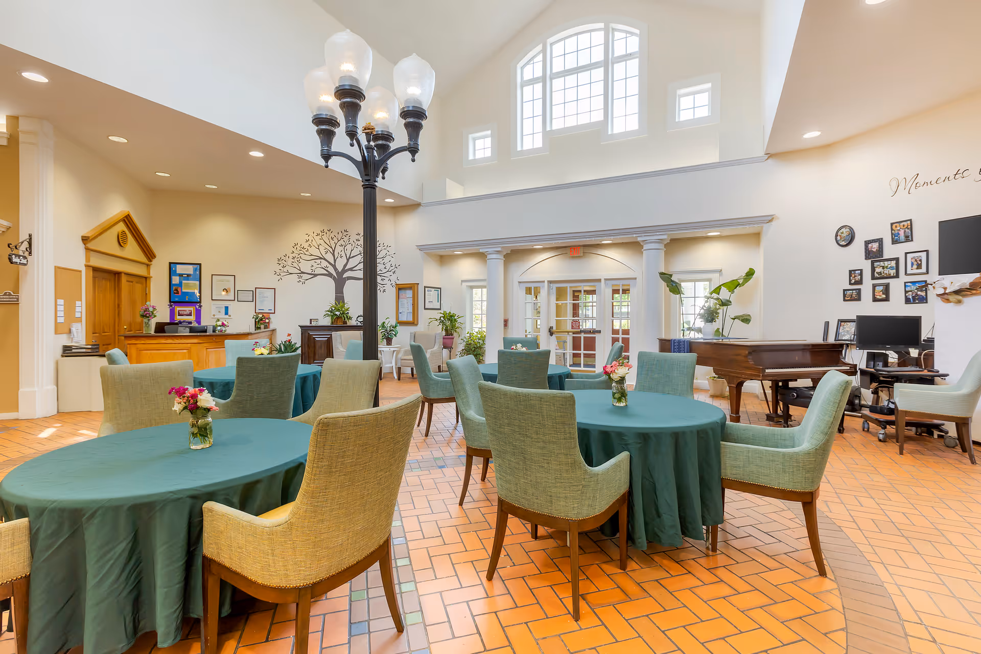 Bright communal dining/lounge area with round tables covered in green tablecloths, upholstered chairs, a grand piano, and a tall arched window.