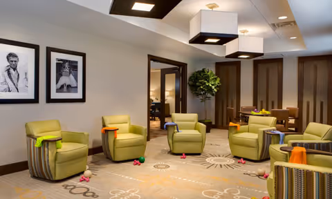 A cozy senior living common area with six green armchairs arranged in a circle on a patterned carpet. Each chair has a colorful cloth draped over the armrest. The room features modern square ceiling lights, framed black and white photos on the wall, and a large potted plant in the corner. A doorway leads to another room with additional seating and a table with fruit.
