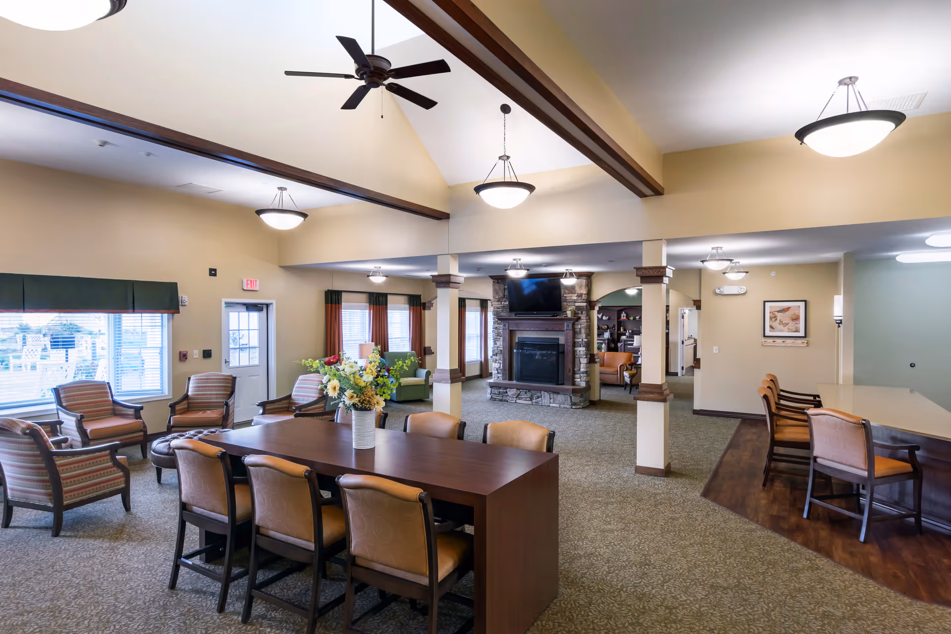 A spacious and well-lit common area in a senior living facility featuring a long wooden table with chairs, a stone fireplace with a mounted TV above it, several armchairs arranged near windows, and a counter with additional seating. The room has high ceilings with exposed wooden beams and multiple ceiling lights.