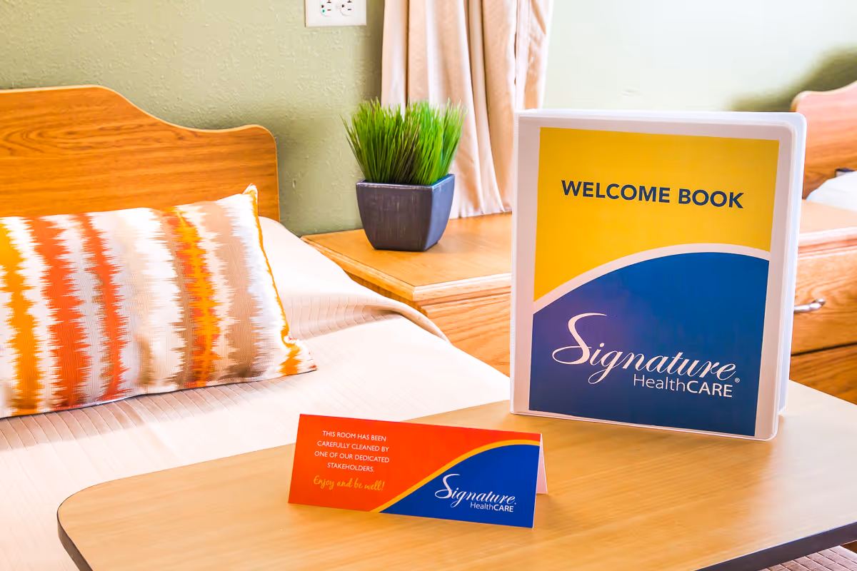 A neatly made bed with a colorful striped pillow and a wooden headboard. On a bedside table next to the bed, there is a small potted plant and a welcome book with the Signature HealthCARE logo. In front of the bed, there is a small card indicating the room has been carefully cleaned by a dedicated stakeholder.