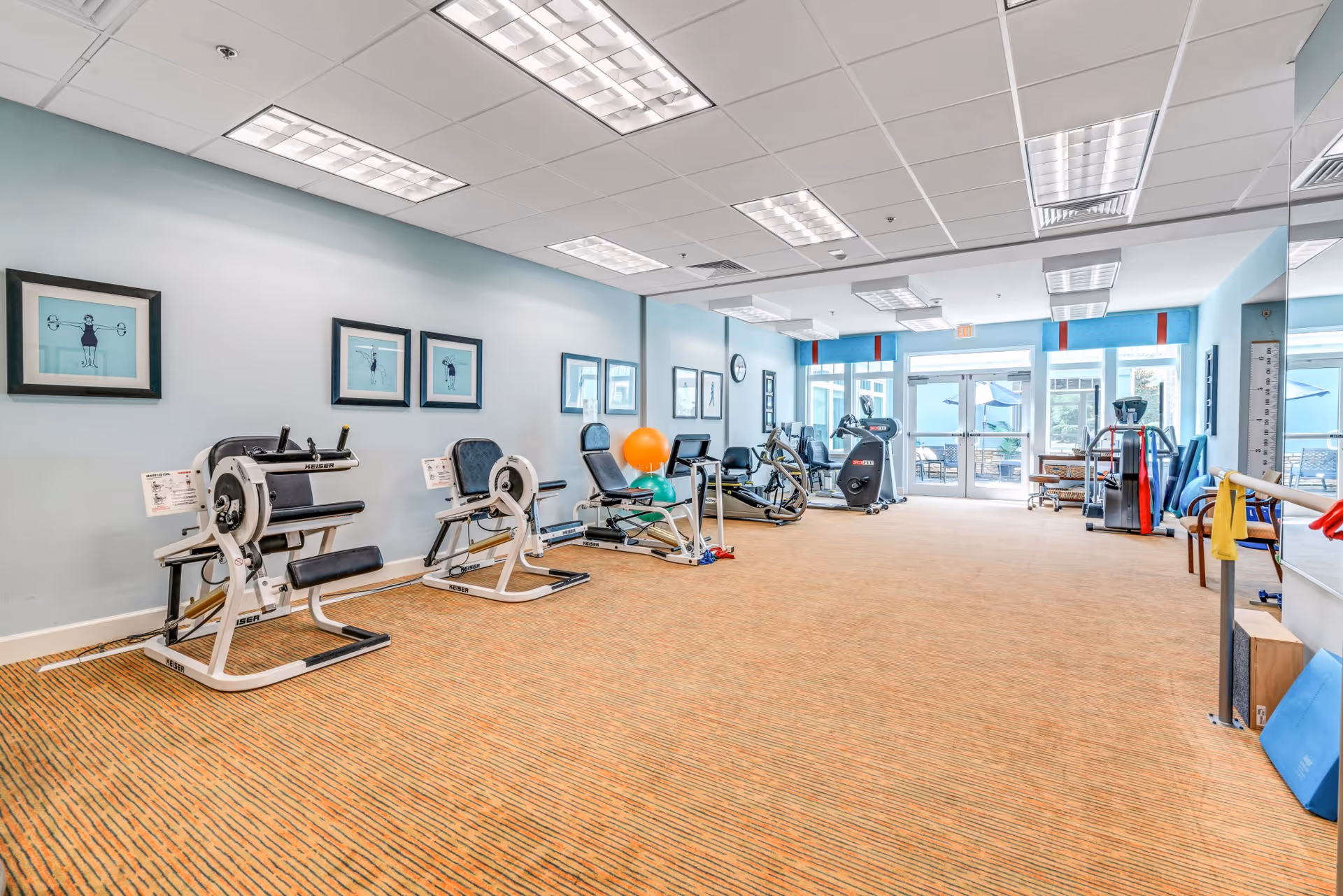 A bright fitness room with exercise machines along the left wall, including seated leg press and rowing machines. The room has light blue walls adorned with framed pictures of people exercising. There are large windows and glass doors at the far end letting in natural light, and exercise balls and resistance bands are visible on the right side near a large mirror.