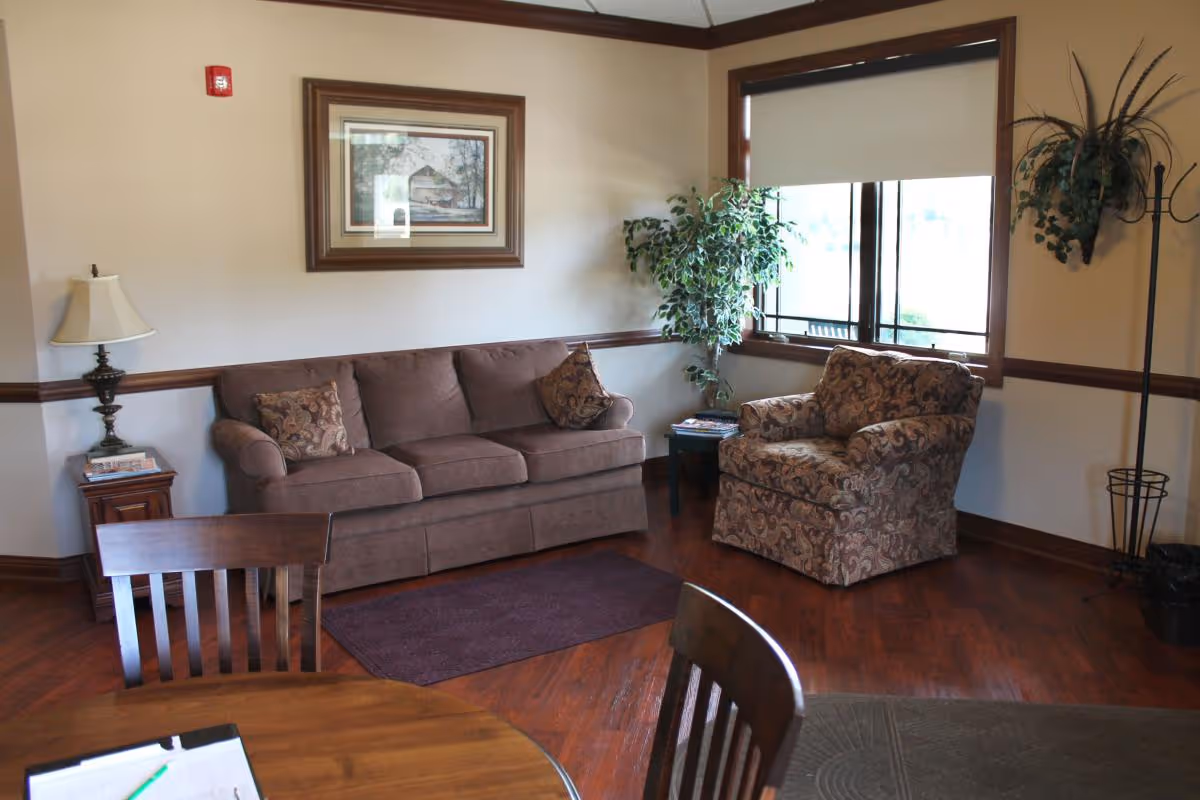 A cozy living room area with a brown sofa, a patterned armchair, a small side table with magazines, a floor lamp, a framed picture on the wall, a large window with a roller shade, a coat rack, and a round wooden table with chairs in the foreground.