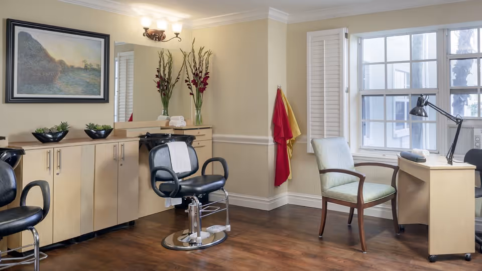 Interior view of a salon area in an assisted living facility with two black salon chairs in front of a long counter with cabinets. There is a large mirror above the counter, a framed painting on the wall, and two vases with tall flowers. A desk with a lamp and a green upholstered chair is positioned near a window with white shutters. Two towels, one red and one yellow, hang on the wall.