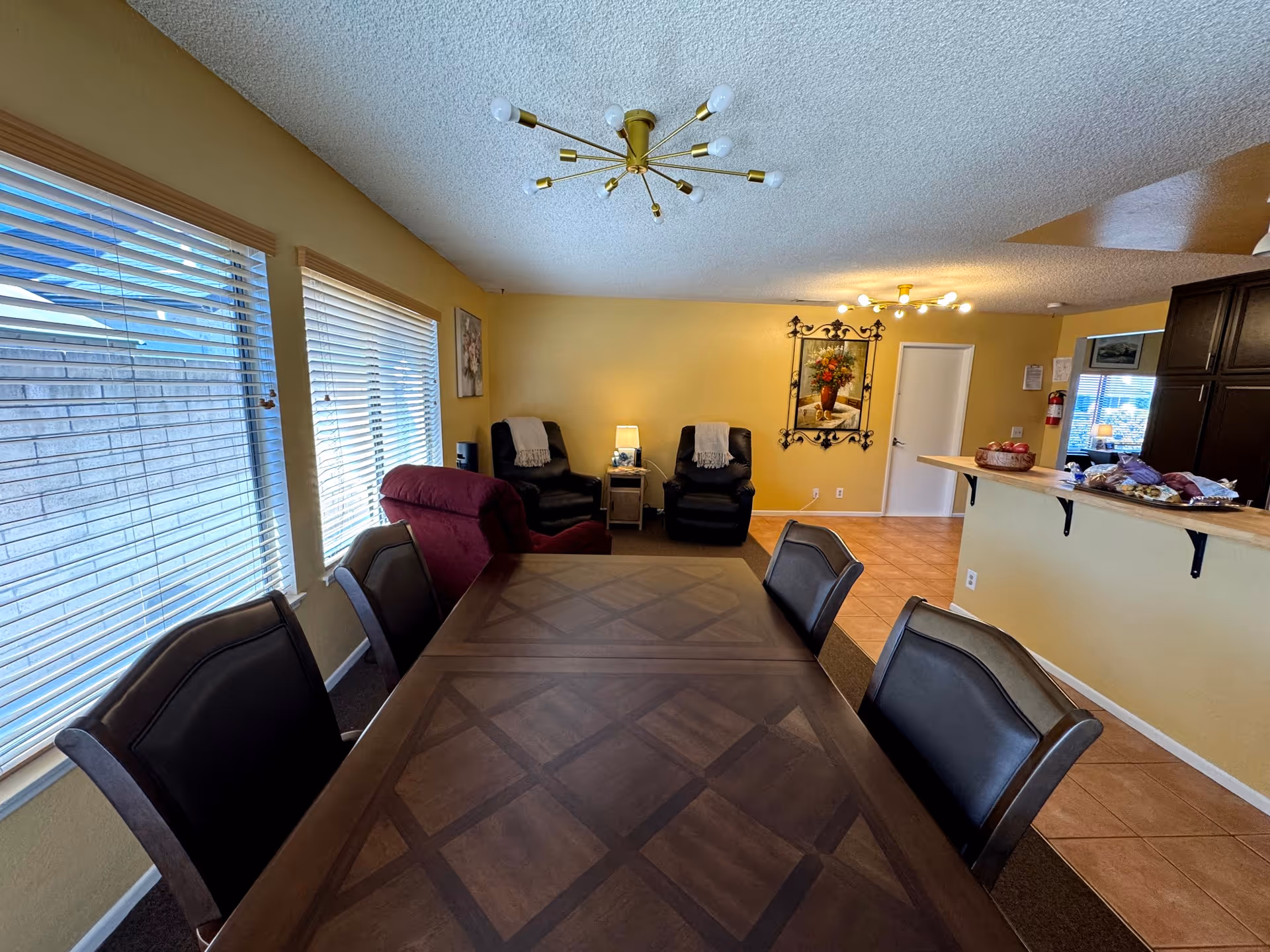 Interior view of a senior care facility room with a large wooden dining table surrounded by six chairs. To the left, there are windows with blinds letting in natural light. In the background, there are two black recliner chairs with a small table and lamp between them, a red recliner chair, and a wall decorated with framed artwork. On the right side, there is a kitchen counter with various items on it and dark wooden cabinets. The walls are painted yellow and the floor is tiled.